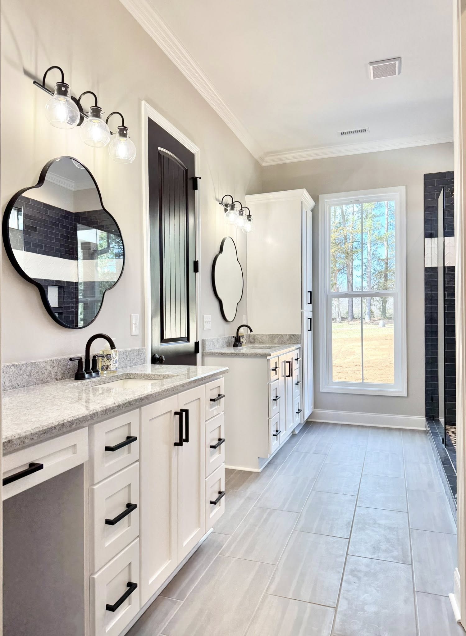 Bright bathroom with dual sinks and white vanities topped with gray granite. Black mirrors and fixtures accentuate the space. A large window offers a scenic view.