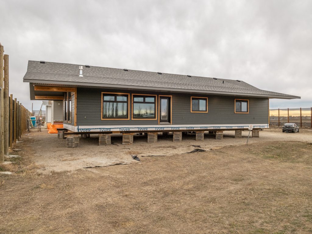 Elevated gray house under overcast sky, supported by wooden blocks, showing large windows and a porch area. The setting is rustic and open.