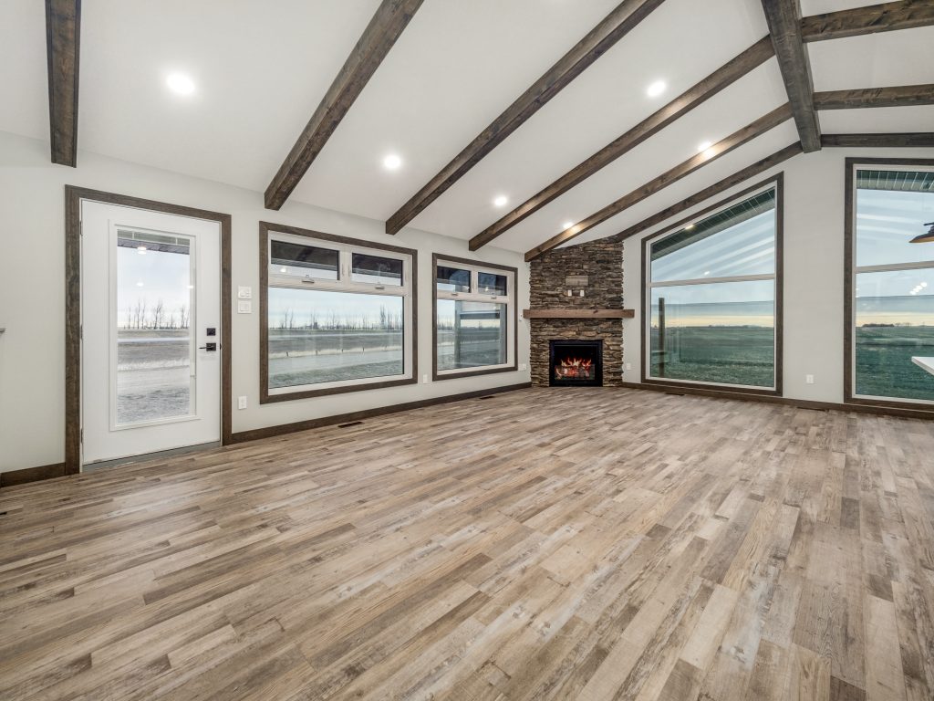 Spacious, bright room with large windows, wood-beamed ceiling, and stone fireplace. Natural light highlights wood flooring, creating a cozy, inviting atmosphere.