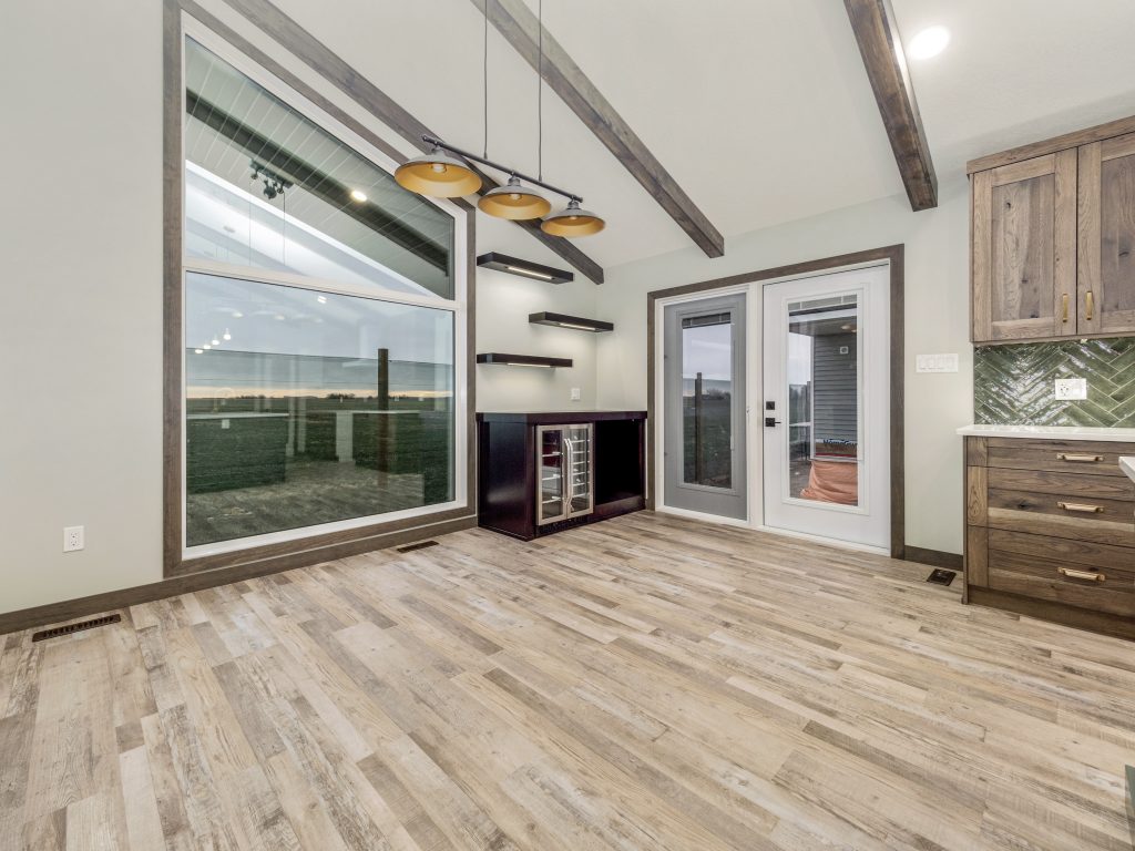 Modern dining room with large windows, a wooden floor, and exposed beams. There are shelves, a wine fridge, and double doors leading outside. Warm, cozy atmosphere.