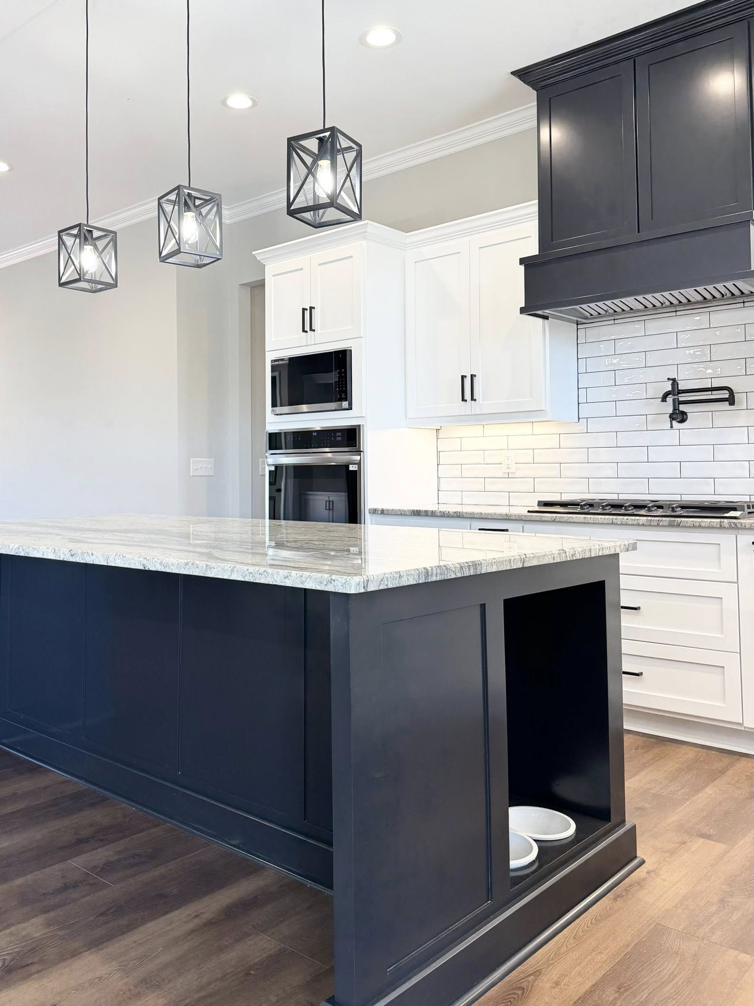 Modern kitchen with a dark island, marble countertop, and pendant lights. White cabinets, tile backsplash, and built-in oven create a sleek, functional space.
