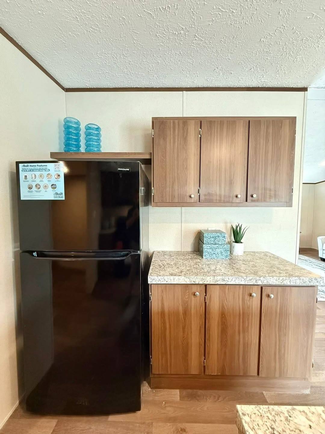 Kitchen corner with a black fridge beside wooden cabinets. Countertop holds a small plant and tissues, and blue glass decor sits atop the fridge. Cozy and tidy.