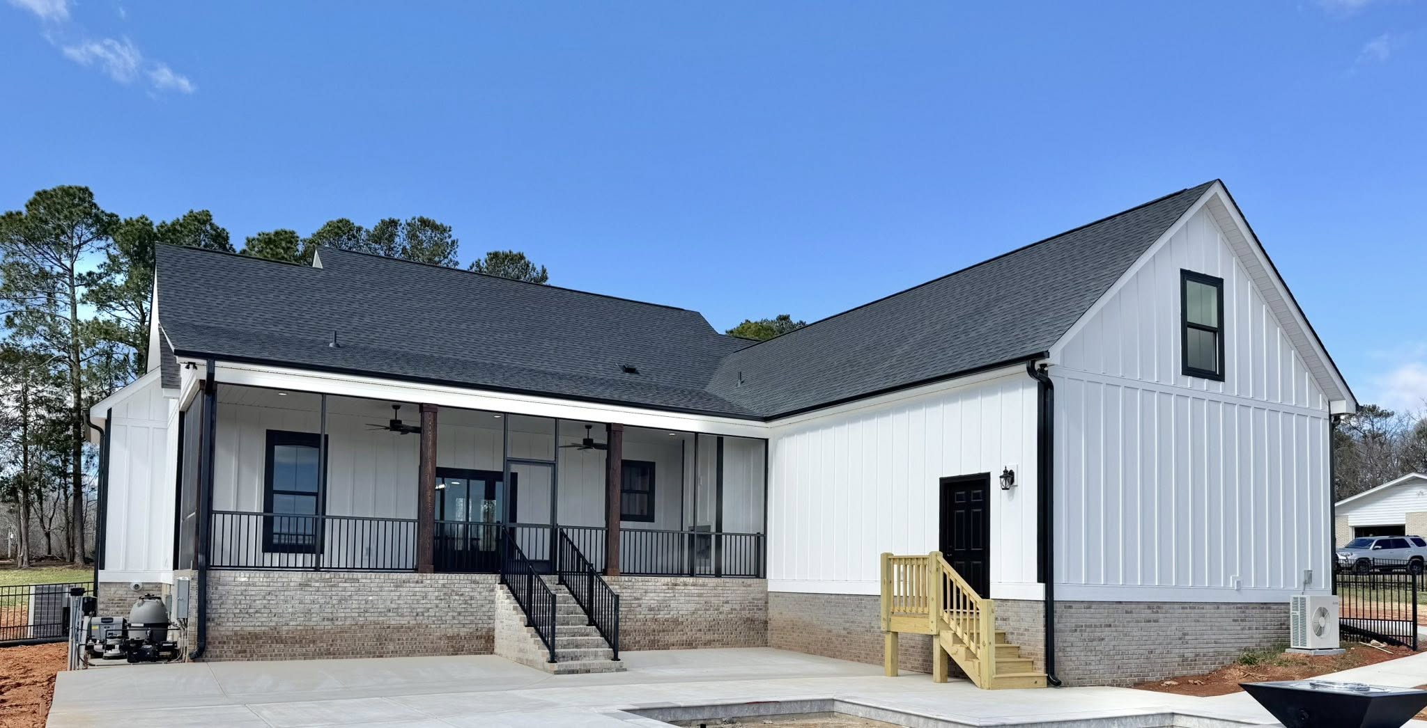 Modern white farmhouse with black roof and porch railings, set against a clear blue sky. Brick foundation, small wooden steps, tranquil and inviting.