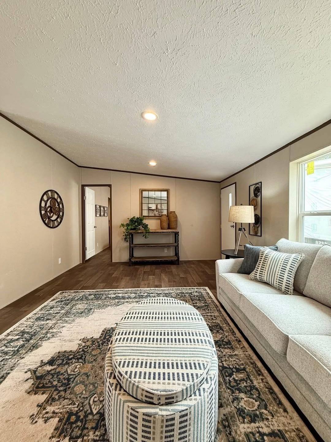 Spacious living room with beige walls and wooden floor, featuring a patterned rug, striped ottoman, gray sofa, and decorative wall art. Warm, welcoming ambiance.
