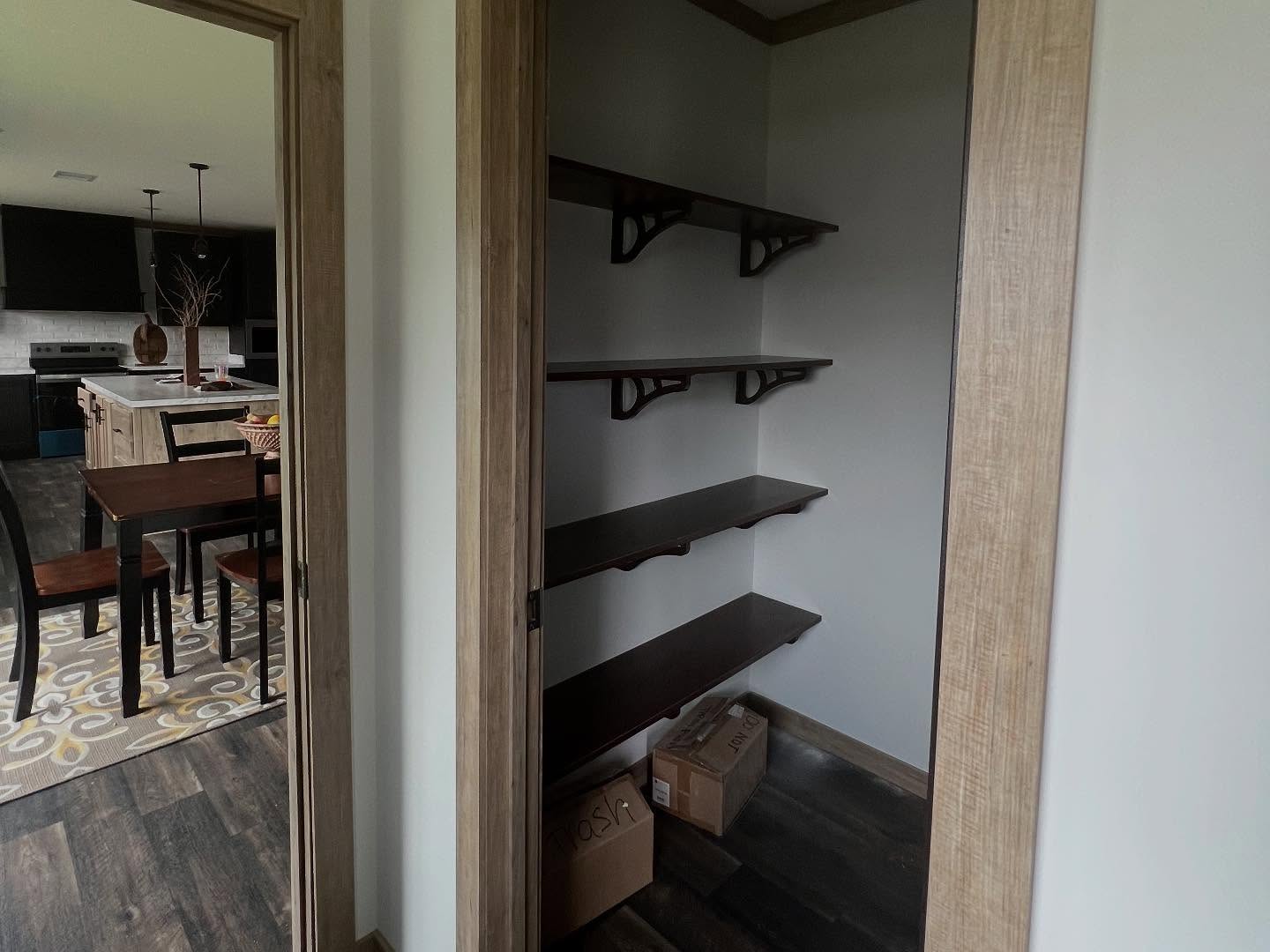 View of an empty pantry with wooden shelves and two cardboard boxes on a dark floor. Dining area visible in the background with a table and chairs.
