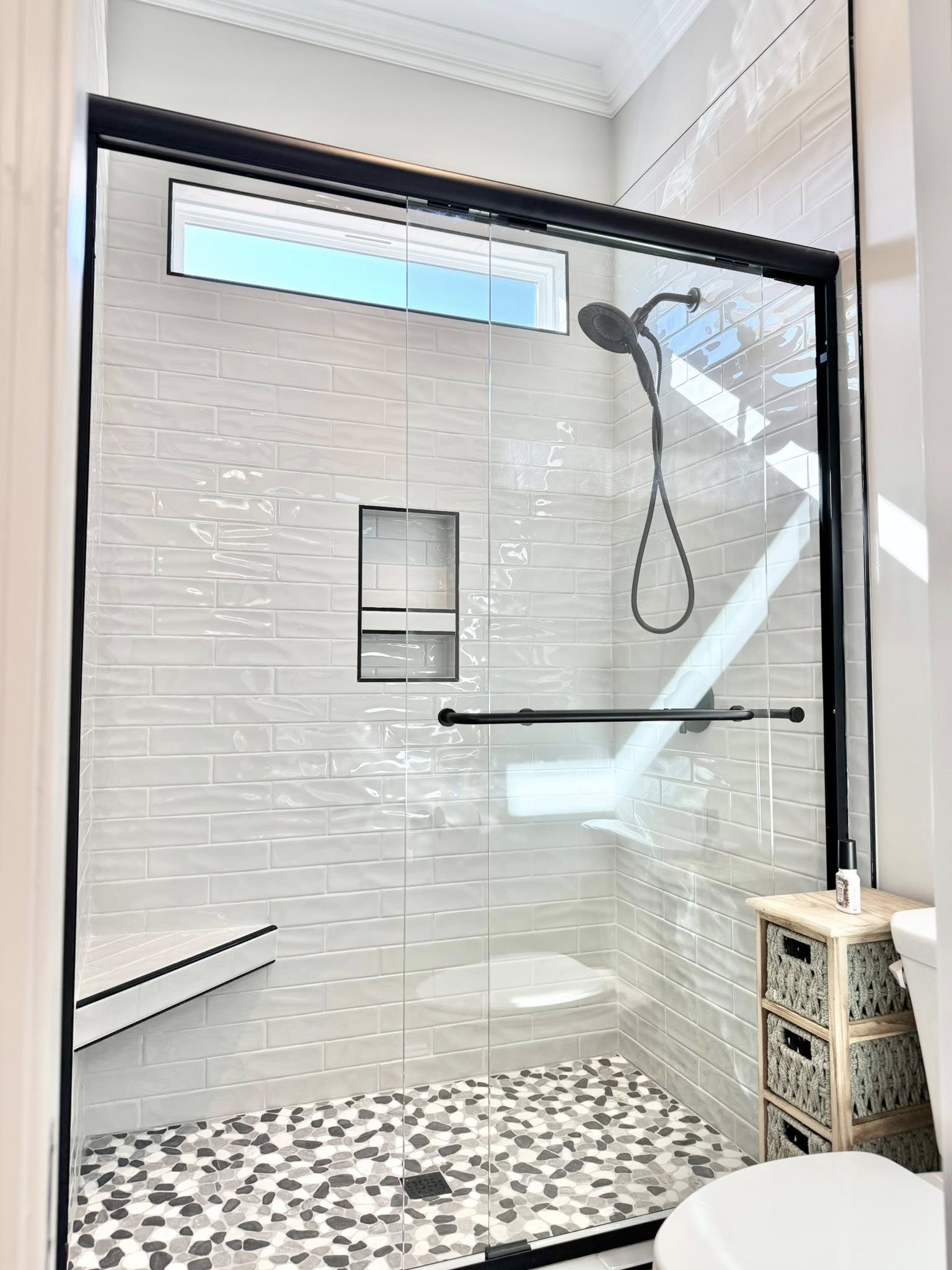 Modern shower with black trim features a rainfall showerhead, white subway tiles, a pebble stone floor, and a glass door. Bright, clean, and minimalist ambiance.