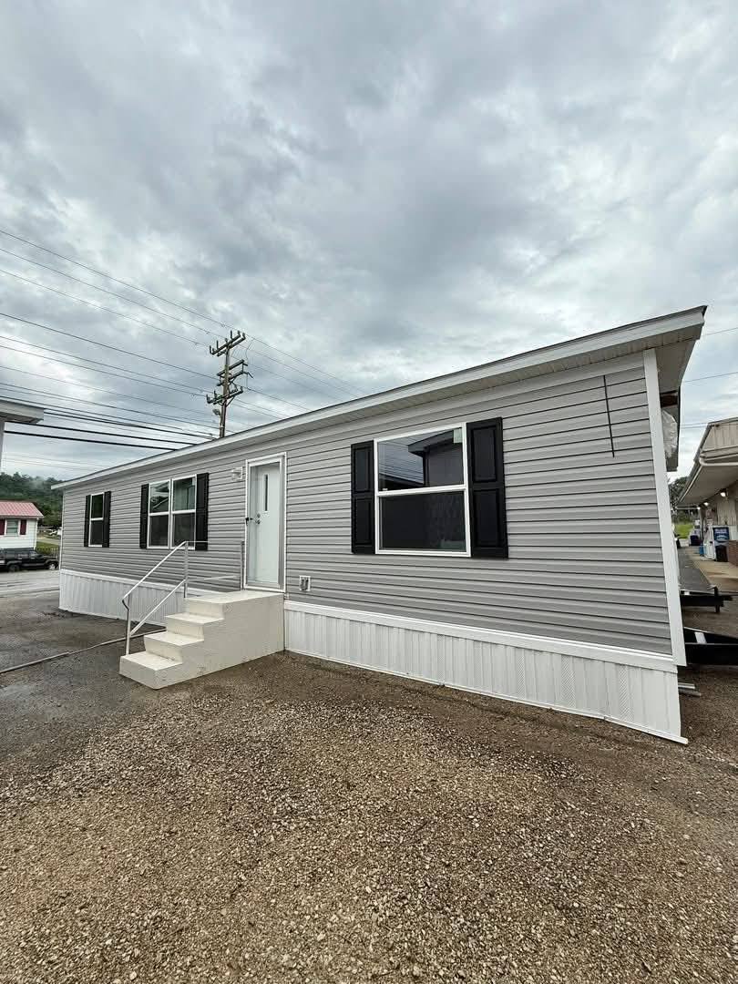 Alt text: "A gray mobile home with black shutters and a white entry door sits under a cloudy sky. Steps lead to the door, and the setting feels calm."