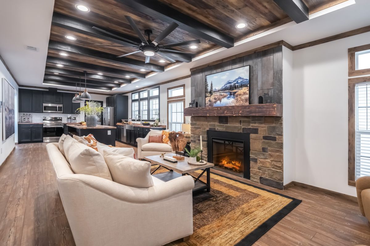 Spacious living area with a cozy stone fireplace, beige sofas, and rustic wood beams. Open kitchen with stainless appliances in the background. Warm, inviting atmosphere.