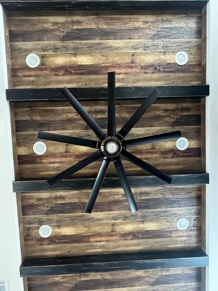 Ceiling with rustic wood panels, featuring a large black ceiling fan in the center and four round white lights evenly spaced around it.