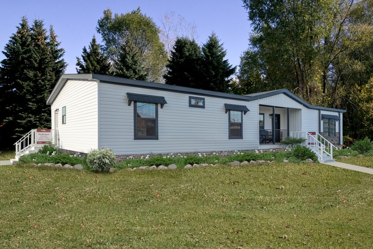 A white manufactured home with a dark roof set on green grass. There's a small stone border with shrubs, and tall trees in the background under a clear sky.