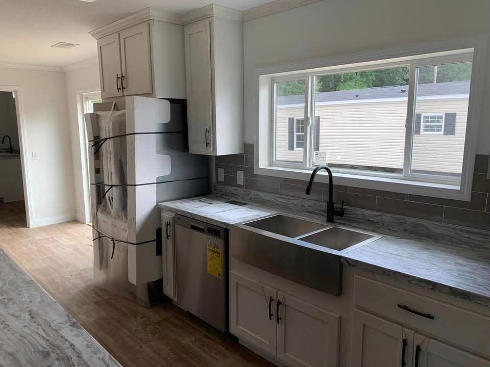 Modern kitchen with light wooden floors, white cabinets, and gray marble countertops. Large window over double sink, and a fridge wrapped in packaging.