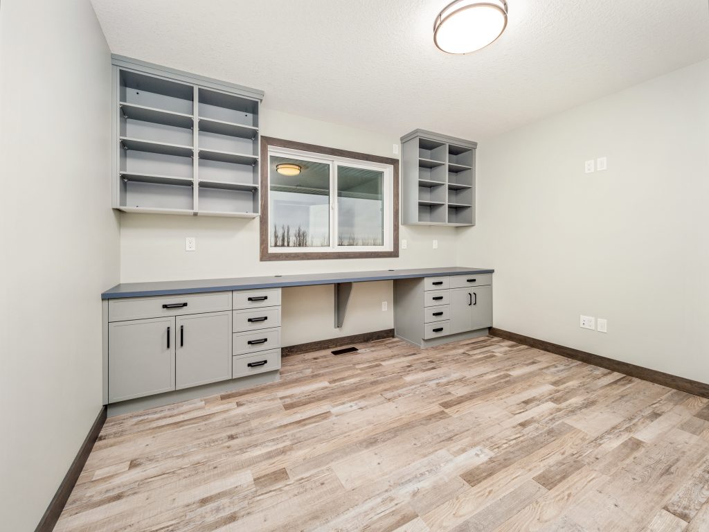 Empty home office with light wood flooring, grey cabinets, and open shelves. A large window lets in natural light, creating a bright atmosphere.