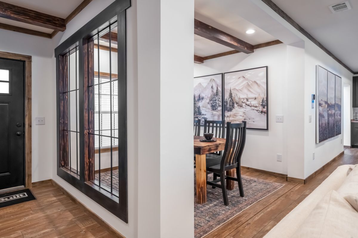 A modern interior features a wooden dining table with black chairs, mountain artwork on white walls, dark wooden beams, and a black front door. Cozy and inviting.