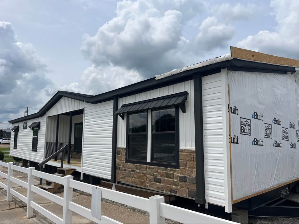 A newly built manufactured home with white siding and stone accents under a cloudy sky. Temporary construction wrap is visible on one end.