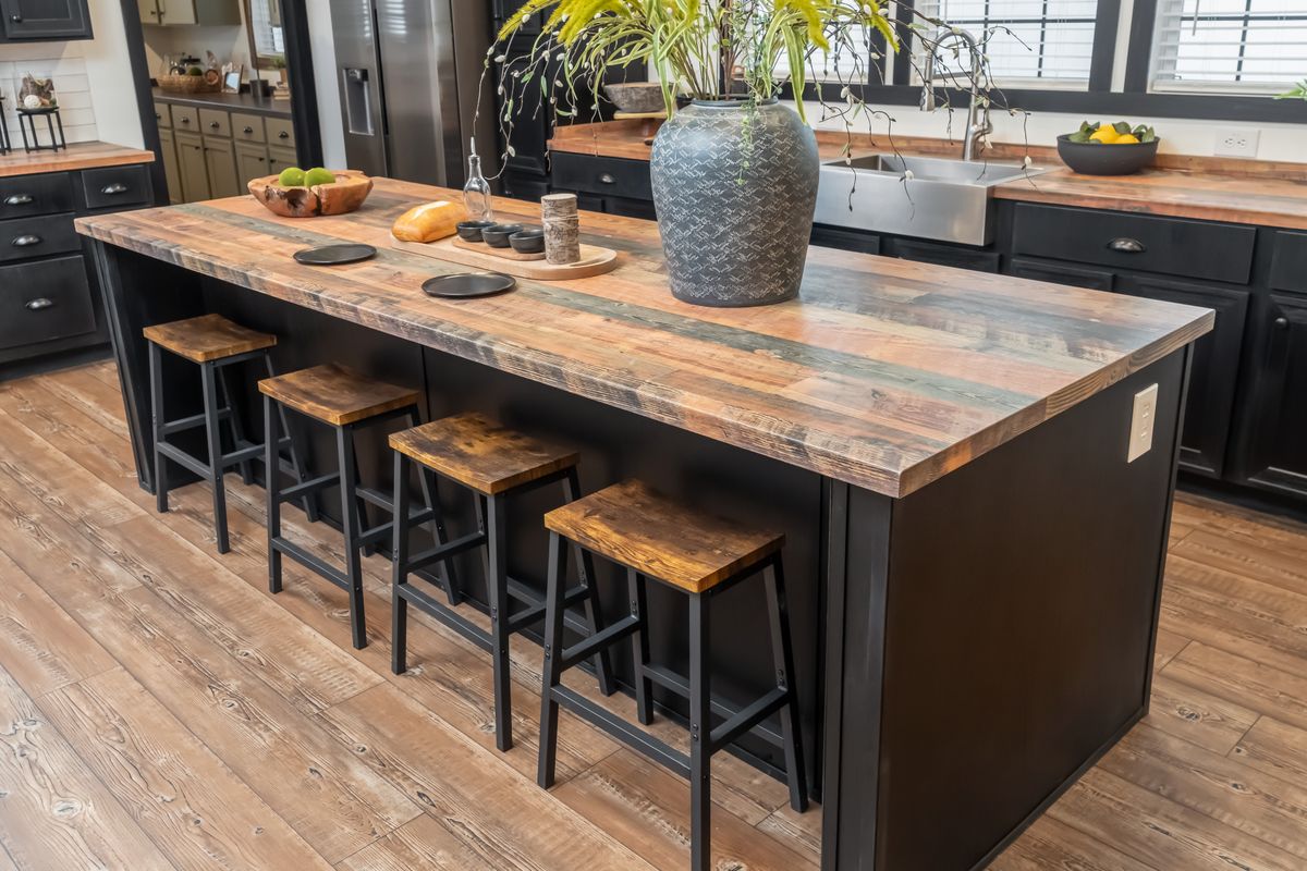 Rustic kitchen with a wooden island, four stools, and a large vase with greenery. Warm tones and natural textures create a cozy, inviting atmosphere.