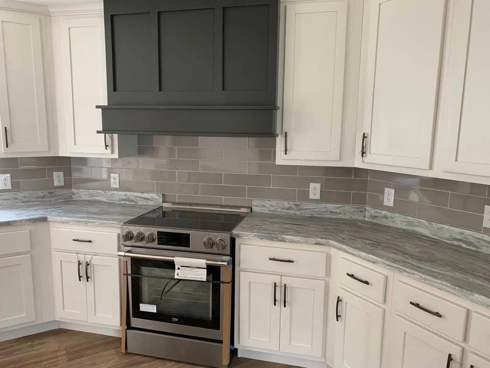 Contemporary kitchen with white cabinets, gray subway tile backsplash, and marble countertops. Centered stainless steel oven under a dark hood.
