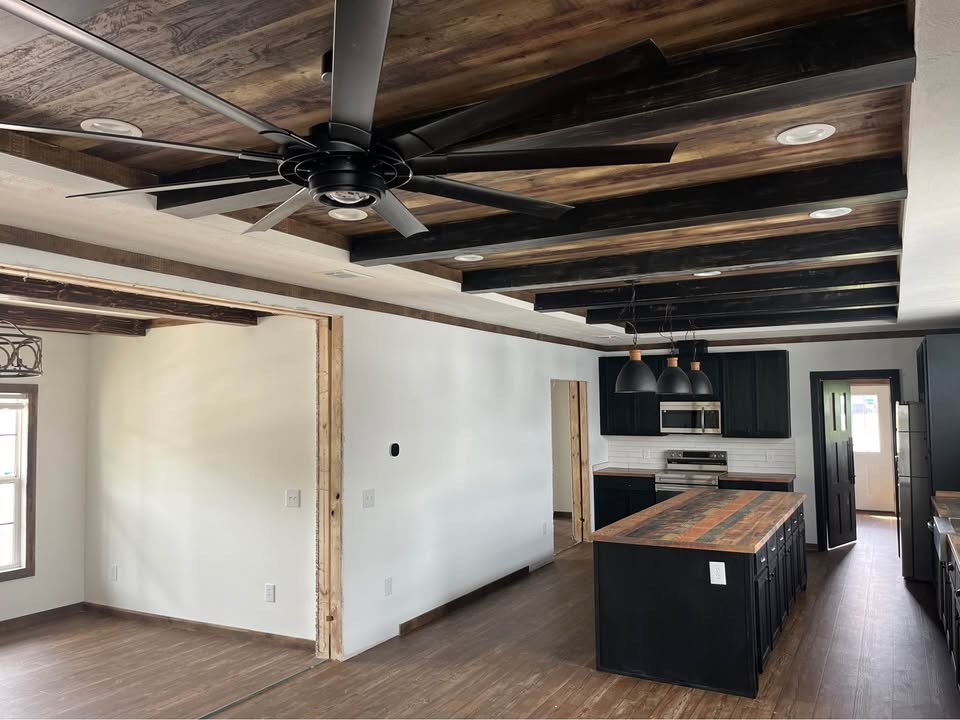 Spacious kitchen with wood-beam ceiling, large black fan, black cabinets, rustic island, and pendant lights. Bright, modern, and inviting atmosphere.