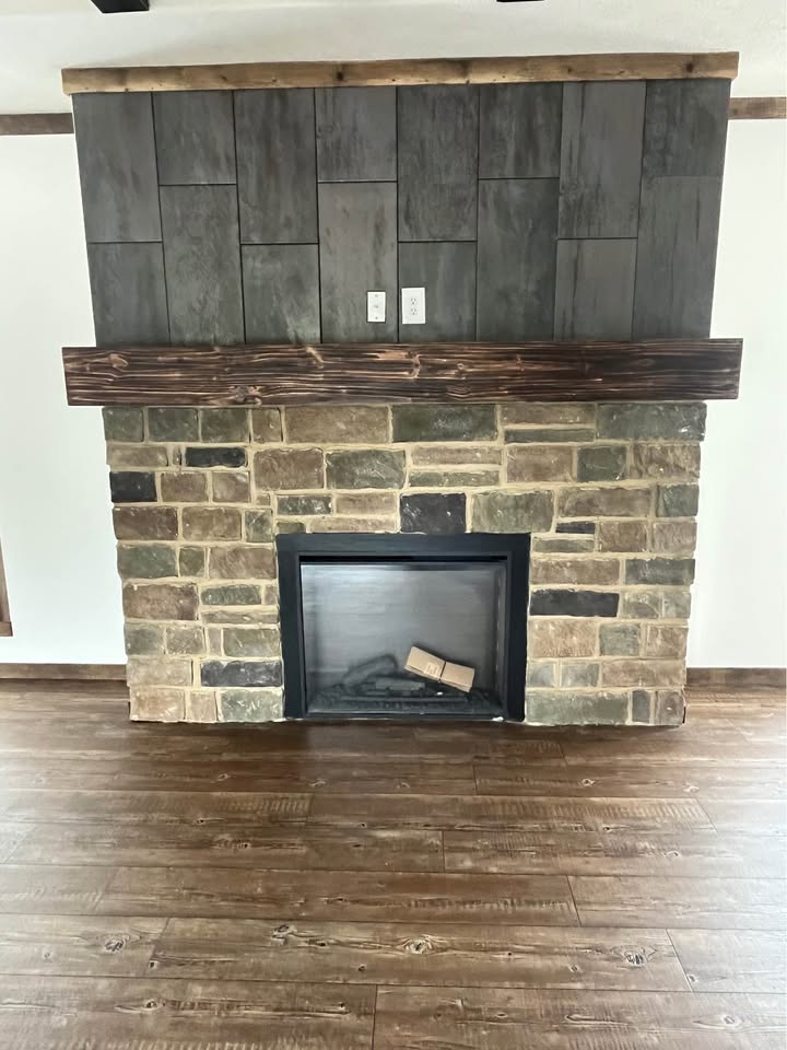 Stone fireplace with a wooden mantel, featuring dark grey tiles above. The hearth is empty, set in a room with rich wood flooring. Cozy ambiance.