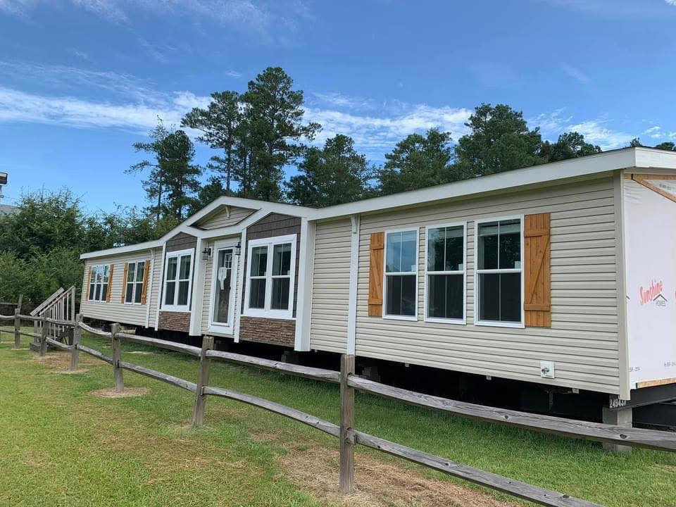 Single-story modular home with beige siding, brown shutters, and a small porch. It's set against a backdrop of trees and a clear sky, creating a serene rural feel.