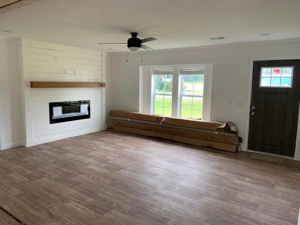 Spacious living room with wooden floors, a ceiling fan, and a large window showing greenery outside. A modern fireplace and dark wood door add style.
