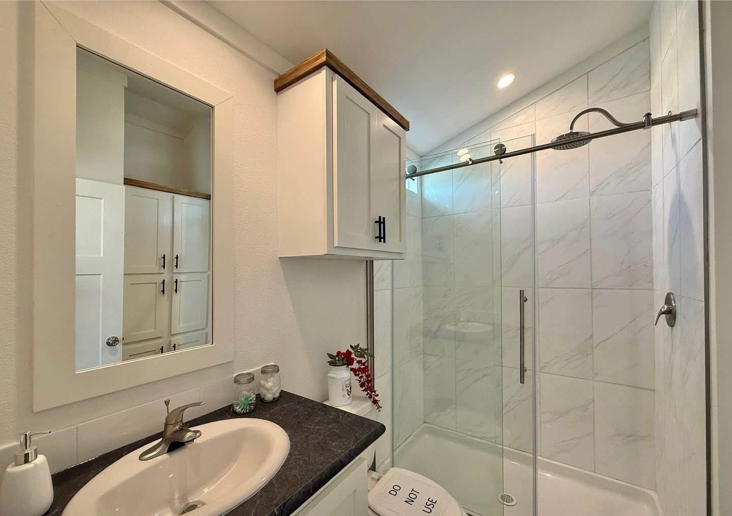 A modern bathroom features a sleek black countertop with a white sink and chrome faucet. Above the sink is a mirrored cabinet. A white cabinet is mounted on the wall, next to a glass-enclosed shower with marble-patterned tiles. The space is bright and minimalist.