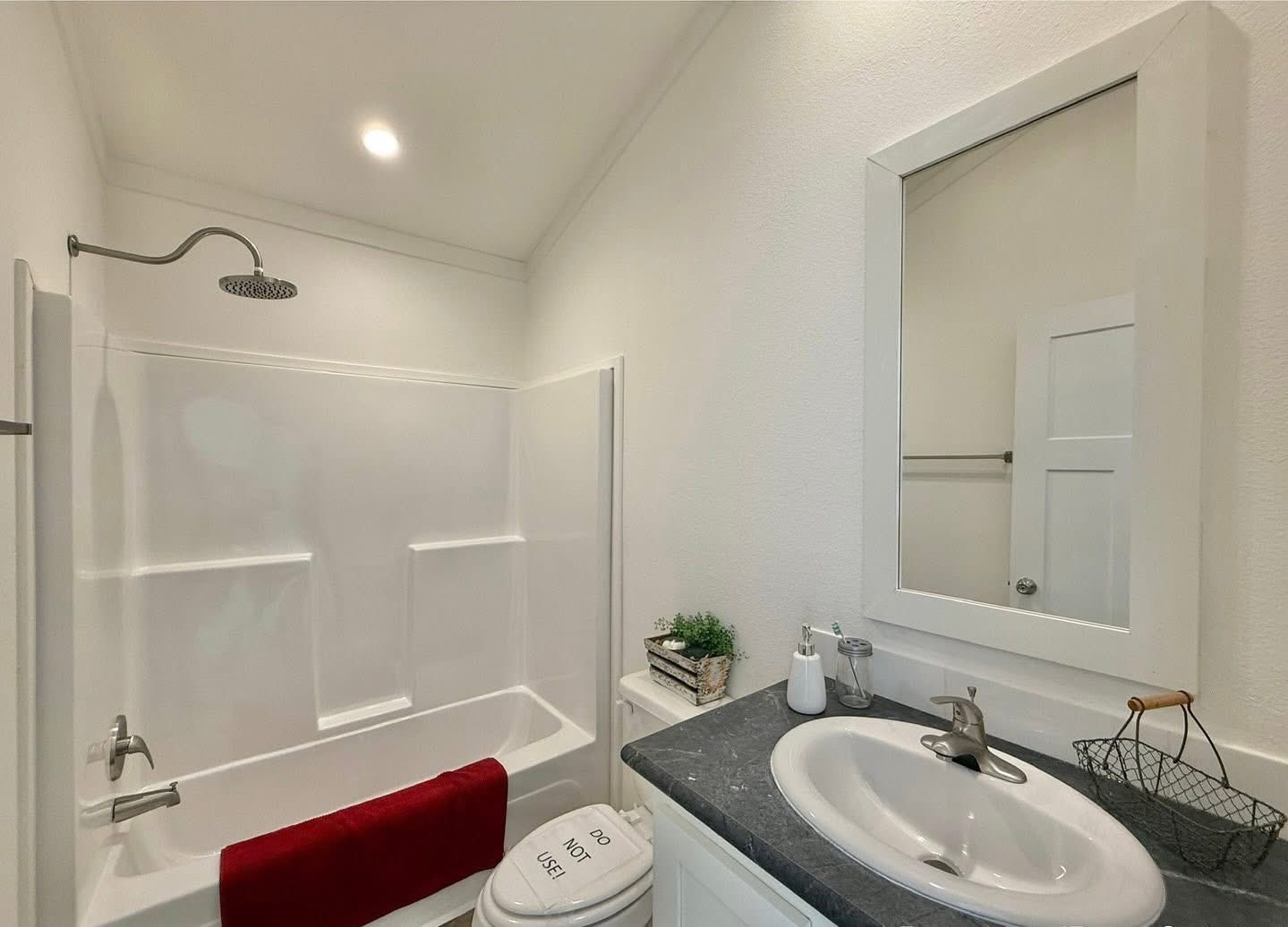 Small, bright bathroom with a white shower-tub, overhead rain showerhead, dark countertop, sleek sink, and red towel; fresh and modern ambiance.