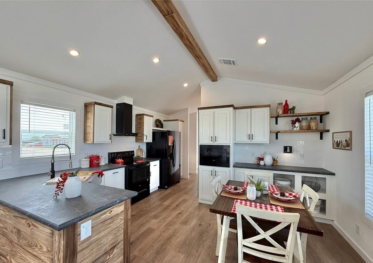 Bright, modern farmhouse kitchen with wooden beams and floors, white cabinets, black appliances, and a dining table set with red checkered accents.