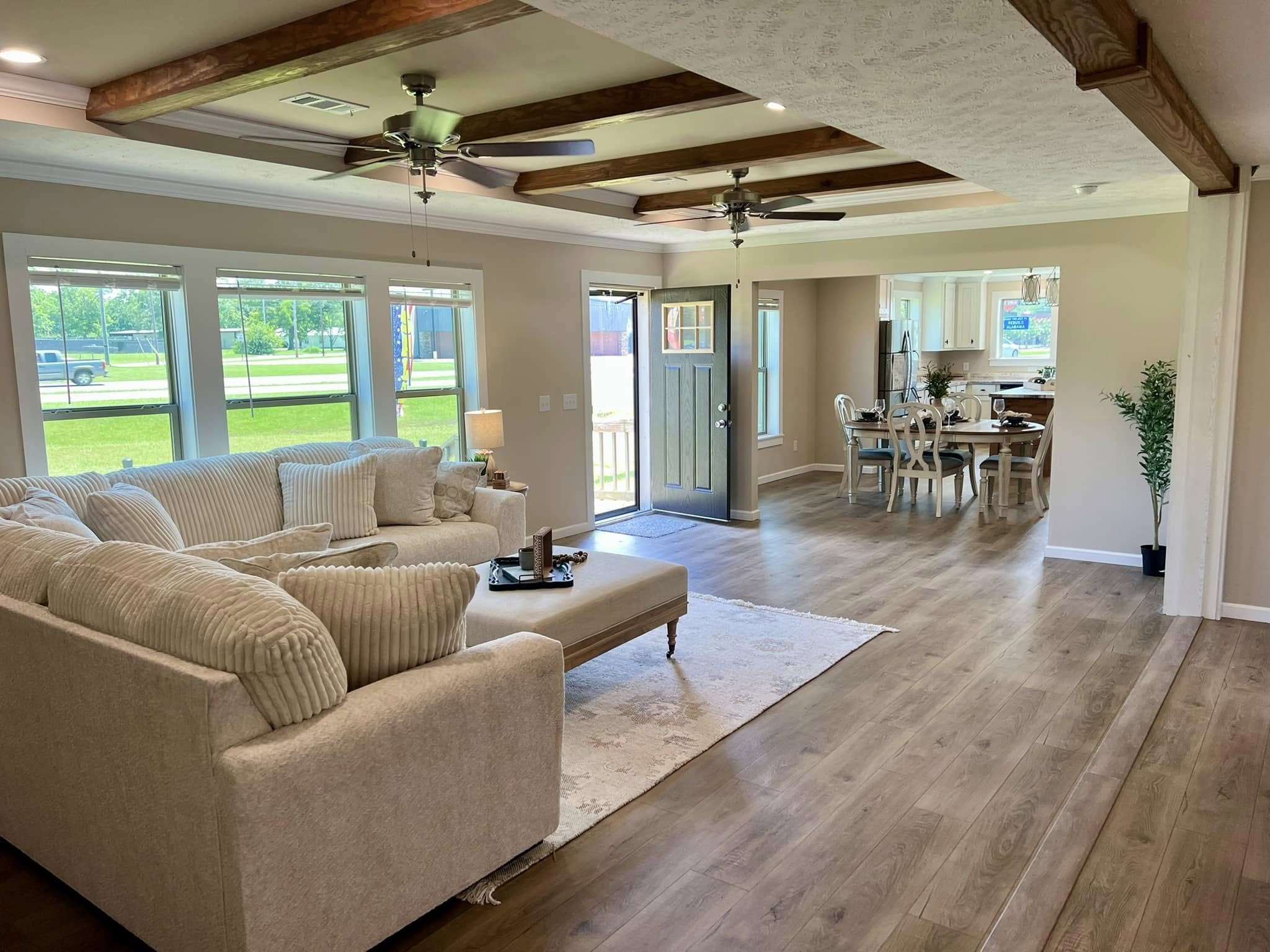 Spacious living area with a light beige sectional, ceiling beams, and wooden floors. Large windows overlook a green yard. Open dining area visible. Cozy, inviting ambiance.