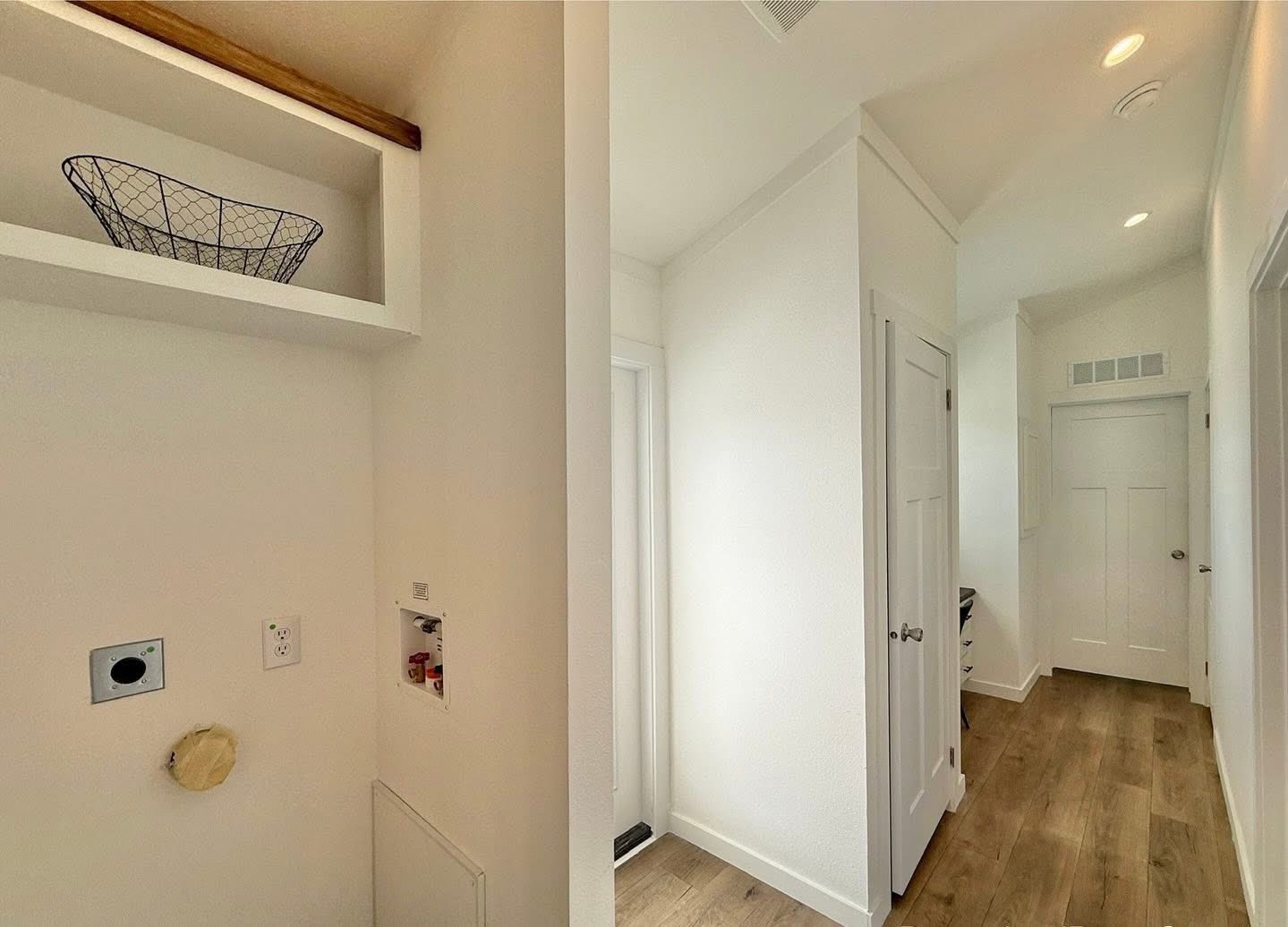 Bright hallway in a modern home with light wood flooring and white walls. Features a laundry area with outlets, shelf, and wire basket on the left.