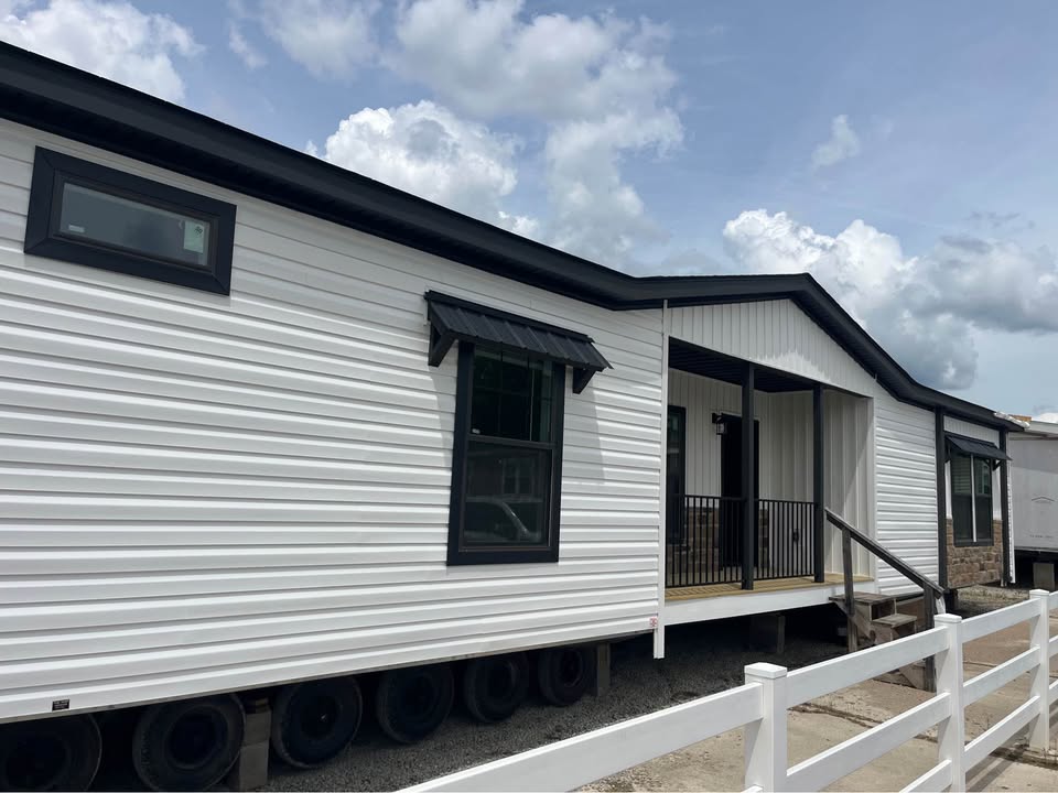 A white manufactured home with black trim is set against a cloudy sky. It features a small porch with steps and a white fence in the foreground.
