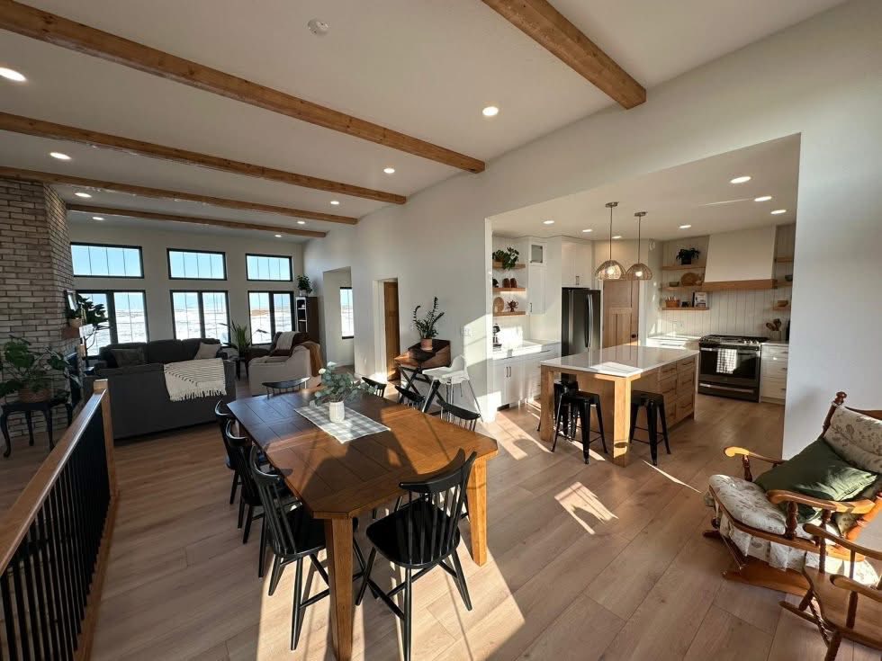 Bright open-concept living space featuring a modern kitchen, wooden dining table with chairs, cozy living area, exposed beams, and large windows. Warm and inviting.