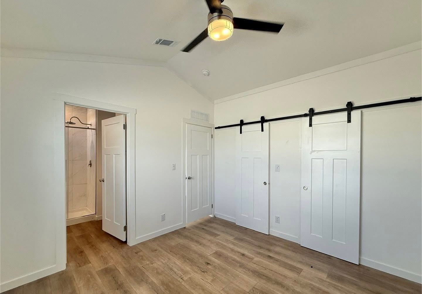 Minimalist bedroom with white walls, wood floor, ceiling fan, two sliding barn doors on the right, and an open door to a shower room on the left.
