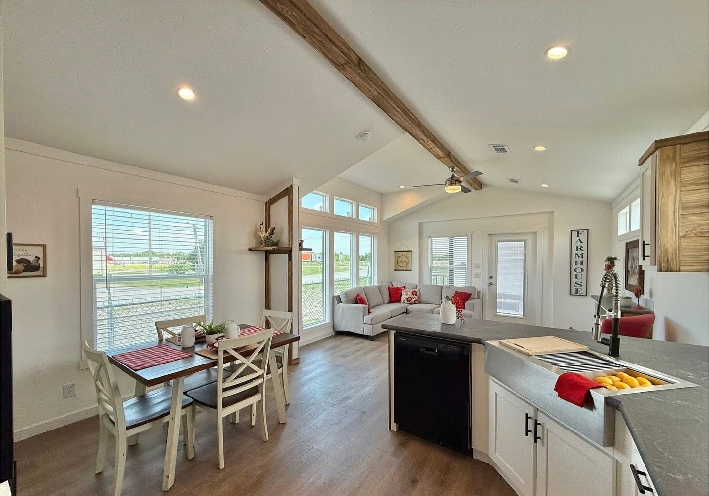Bright, modern farmhouse interior with open layout. Features include a dining table with red accents, cozy sofa, kitchen island, and wood beam ceiling.