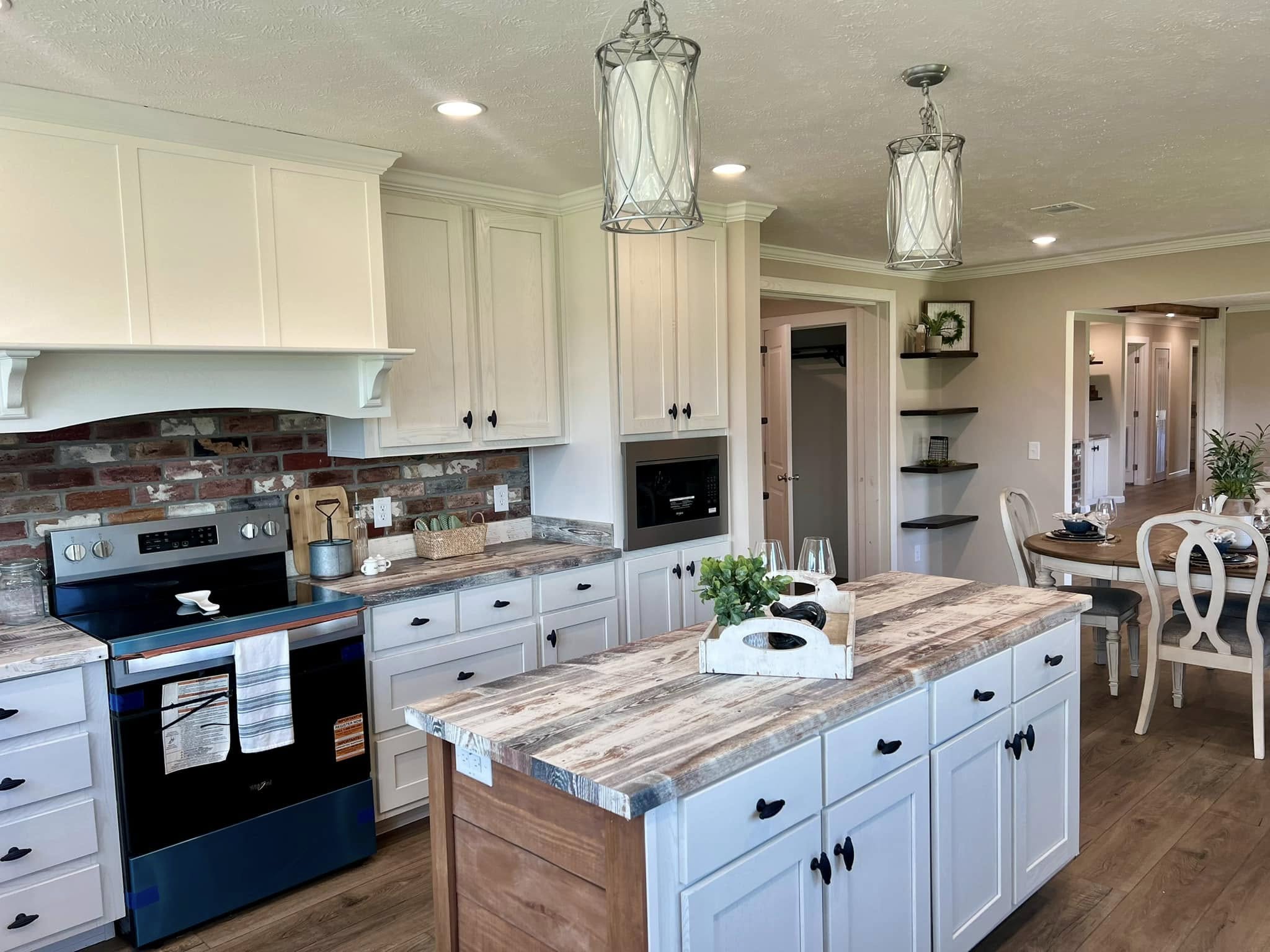 A modern kitchen with rustic charm features white cabinetry, a wooden island, and a brick backsplash. Pendant lights hang above, and a dining area is visible in the background.