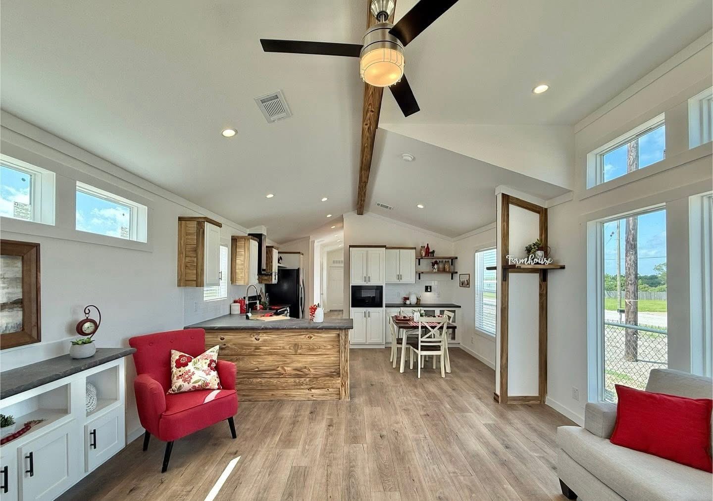 Bright, cozy living space with wood accents, featuring a kitchen island, dining area, red armchair, ceiling fan, and large windows with countryside views.