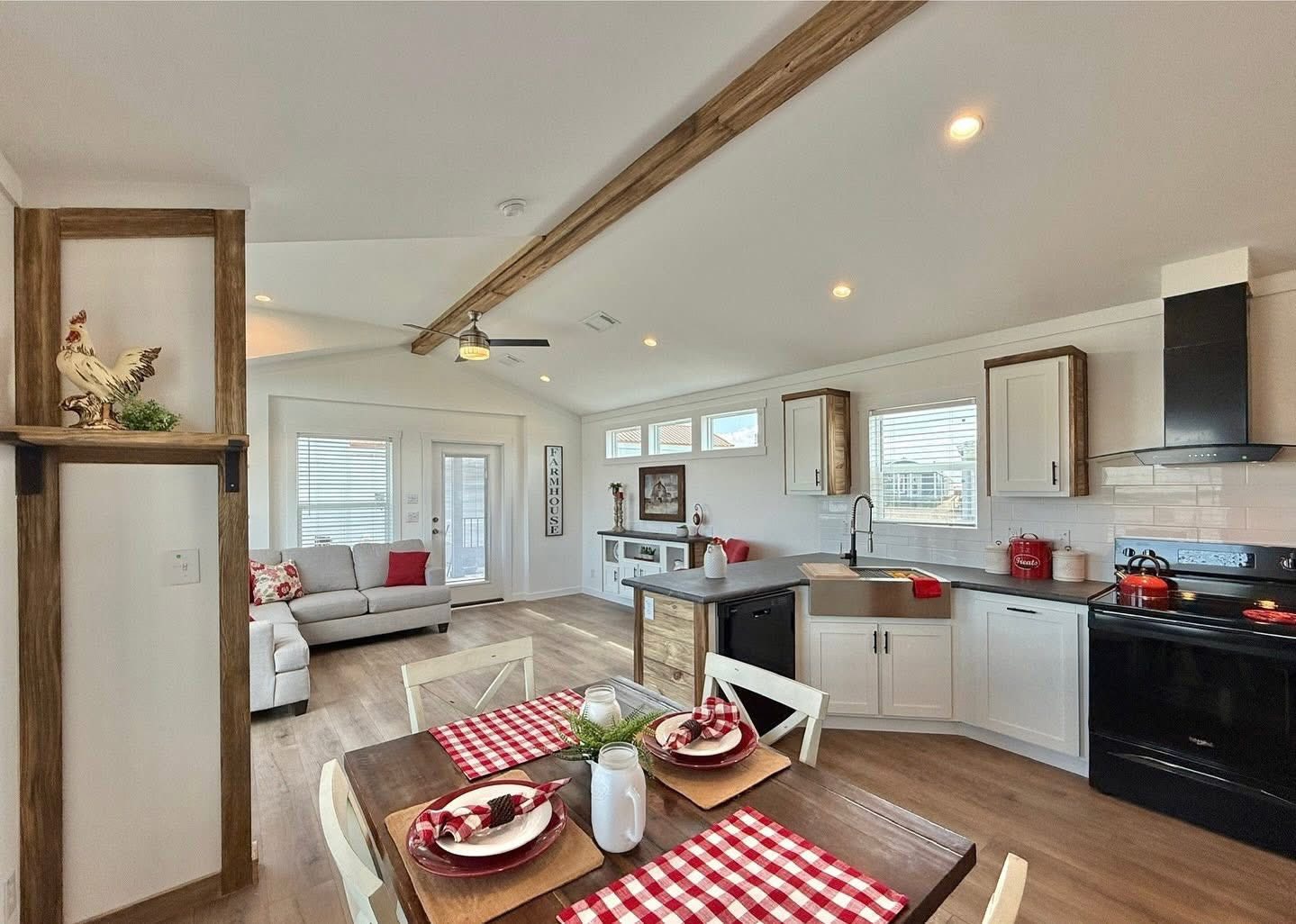 Bright, rustic kitchen-dining area with wood accents. Features a checkered dining table, white cabinetry, and cozy living room with a gray sofa. Warm and inviting.