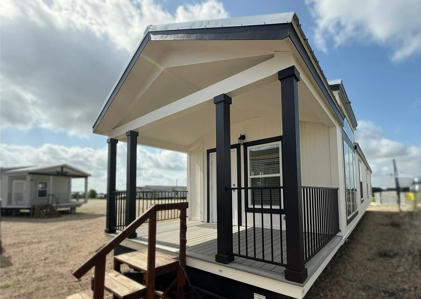 A modern tiny house with a gabled roof and a small porch with black railings. The sky is partly cloudy, creating a bright and open atmosphere.
