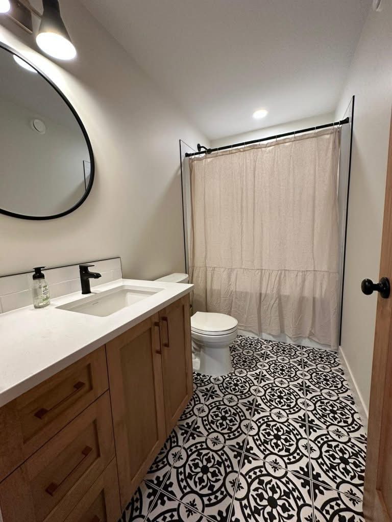A modern bathroom featuring a wooden vanity with a white countertop and black faucet, patterned black-and-white tile floor, and beige shower curtain.