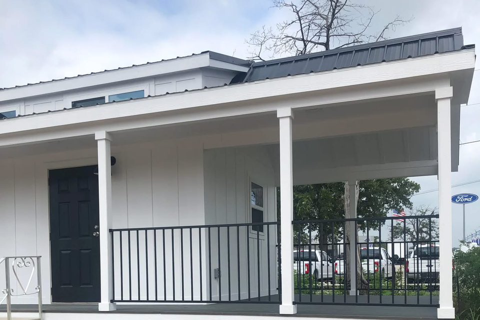 A small white house with a gray metal roof and black door features a porch with black railings. A Ford sign and parked trucks are visible in the background.