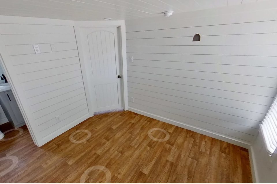 A small, white room with shiplap walls and a wooden floor. Light streams in through window blinds, highlighting the minimalist, clean space.