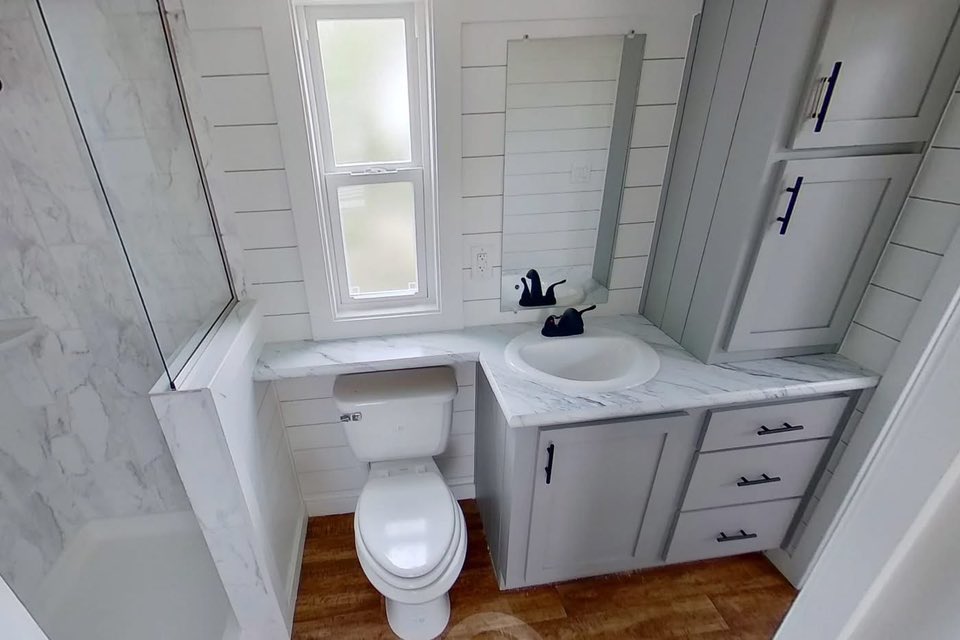 Compact bathroom with white shiplap walls, featuring a toilet and sink with a black faucet on marble countertop. Cabinetry in a light gray tone.
