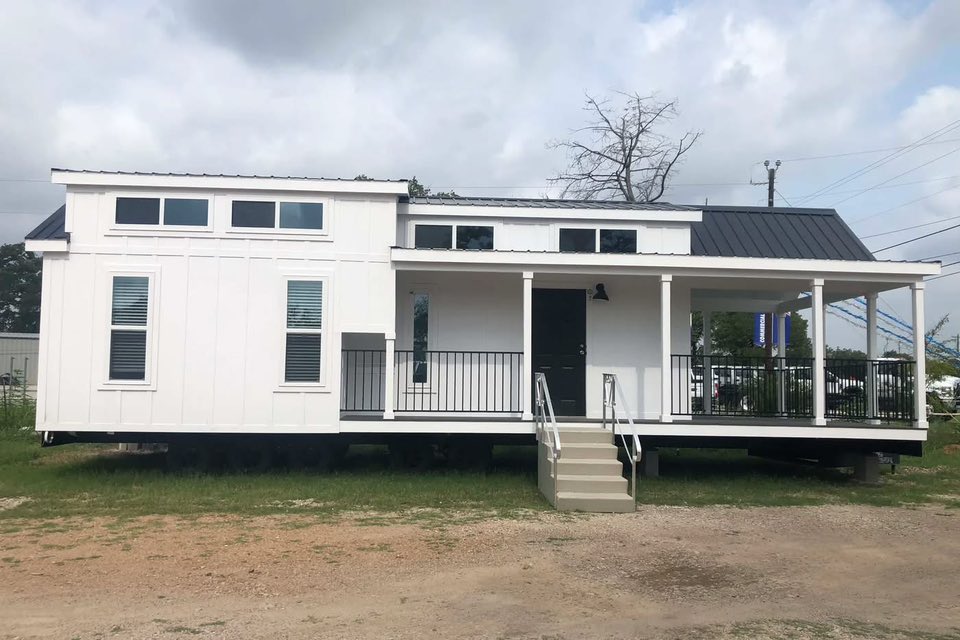 A modern, white tiny house with large windows and a black roof, featuring a covered front porch and steps leading to the entrance. Overcast sky above.