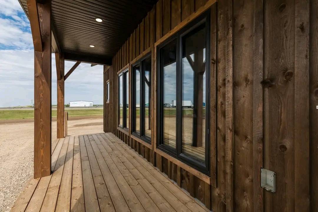 Rustic wooden porch with black-framed windows, overlooking a rural landscape under a partly cloudy sky. Warm and inviting tone.