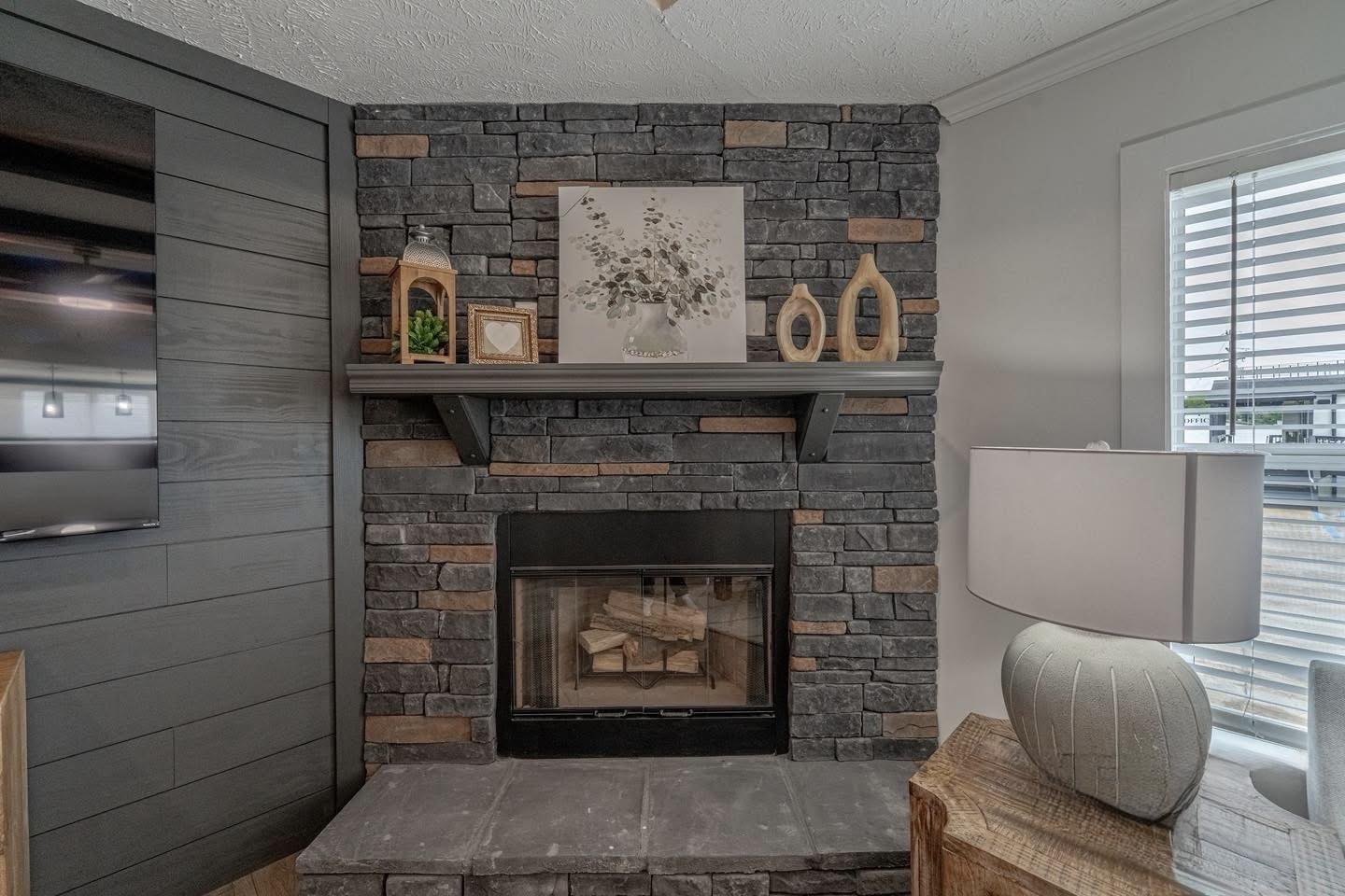 Cozy living room with a dark stone fireplace, decorated with a painting, lanterns, and sculptures on the mantel. A modern lamp sits on a wooden table.