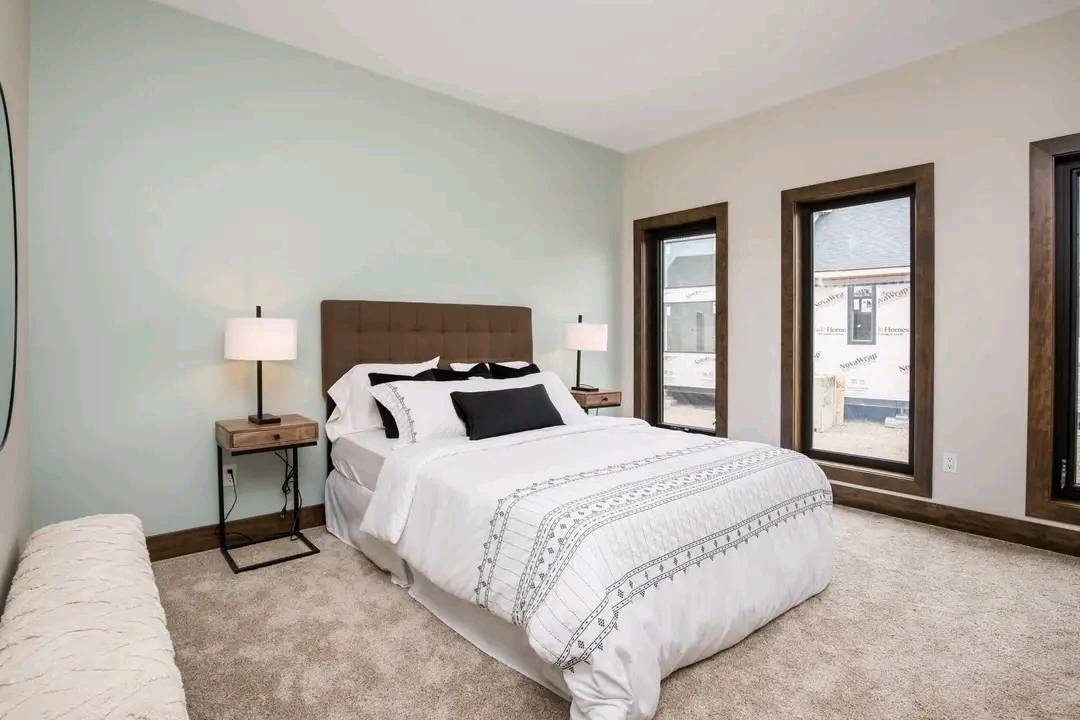A cozy bedroom features a large bed with white and patterned bedding, flanked by two wooden nightstands with lamps. Three tall windows on the right let in natural light, creating an inviting and peaceful atmosphere.