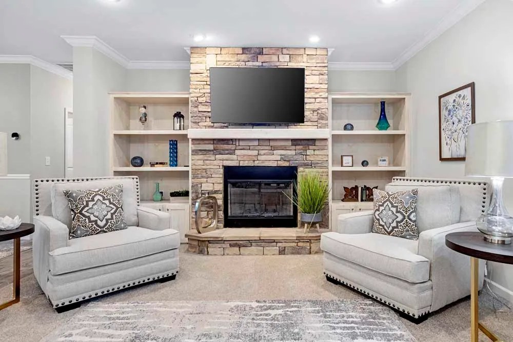 A cozy living room with two beige armchairs facing a stone fireplace topped by a TV. Built-in shelves, soft lighting, and decorative accents create an inviting atmosphere.