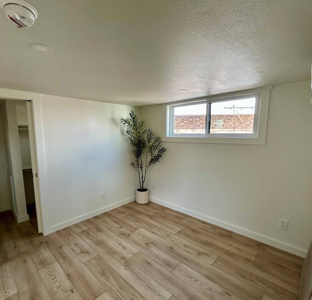 A small, bright room with light wood flooring, white walls, and a ceiling with recessed lighting. A potted plant stands by a low window, creating a minimalist, peaceful vibe.