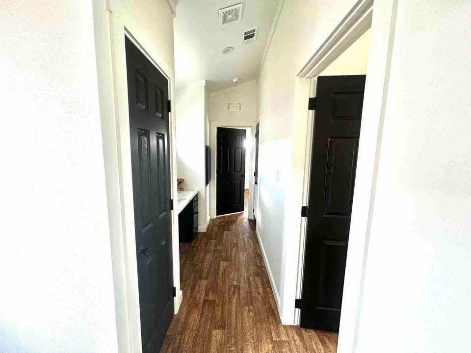 A narrow hallway with wooden floors and white walls features several black doors on either side, leading to different rooms. Bright lighting creates a clean, inviting atmosphere.