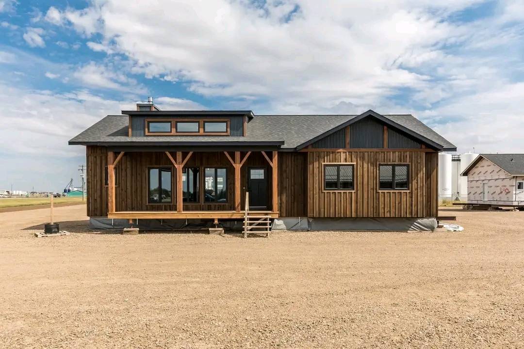A rustic wooden house with a porch and large windows sits on a dirt lot under a partly cloudy sky. The scene conveys a sense of simplicity and tranquility.