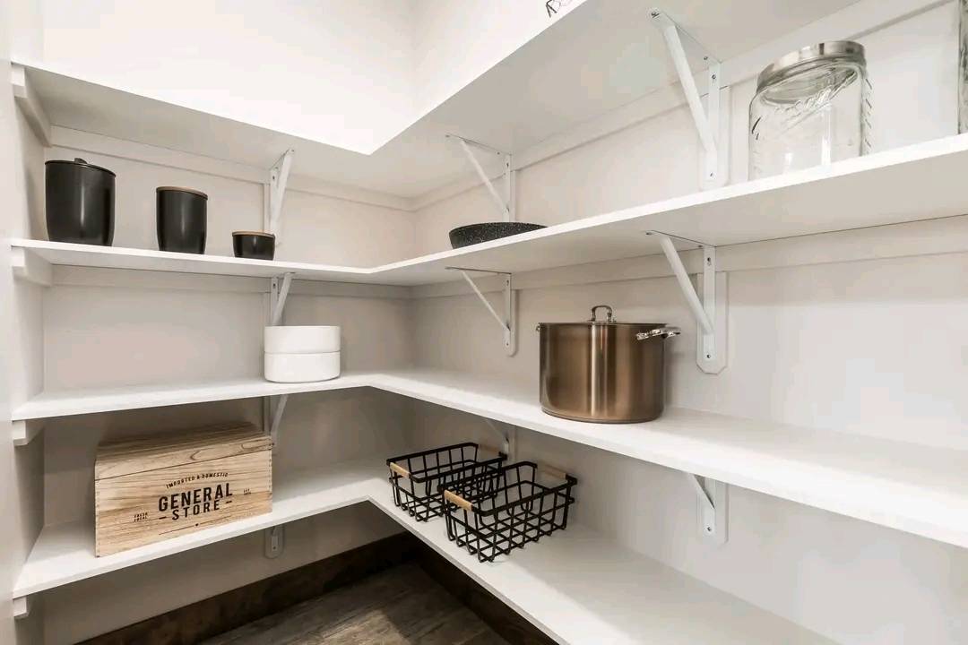 A well-organized pantry with white shelves featuring black containers, a glass jar, a metal pot, wire baskets, and a wooden box, conveying neatness.